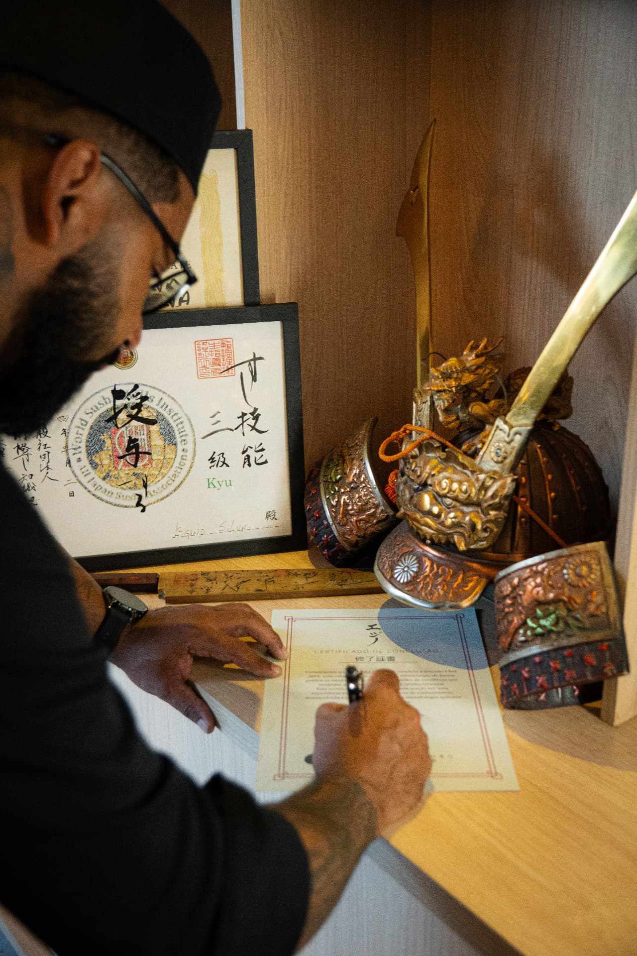 Chef Egino Silva assinando um certificado ao lado do diploma do World Sushi Skills Institute e peças japonesas do instituto Kenkyo