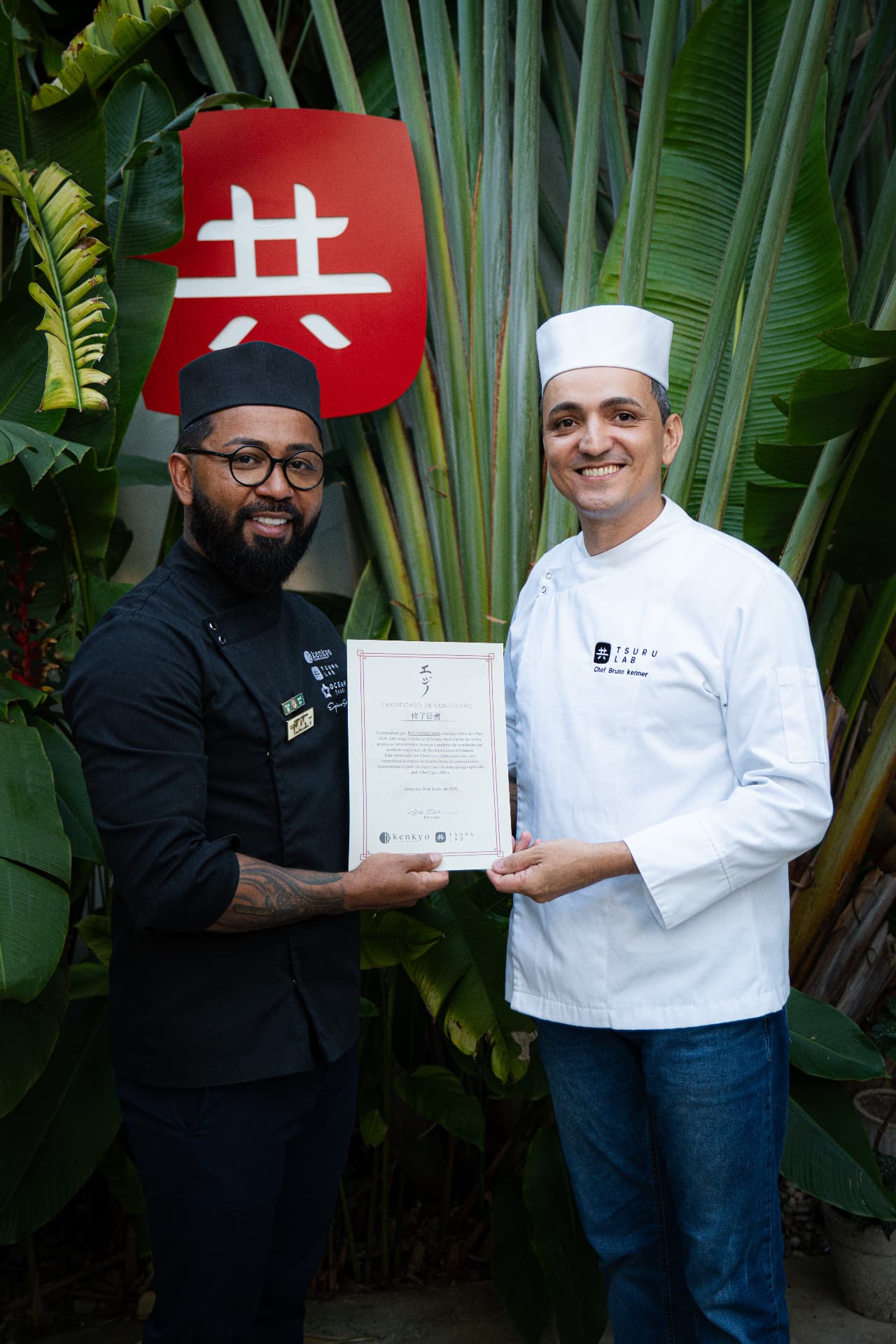 Chef consultor Egino Silva recebendo o certificado de conclusão das mãos do chef Bruno Kenner, do TSURU LAB, ao lado do símbolo Kenkyo